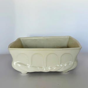 May include: Cream-colored rectangular ceramic planter with a decorative arched design along the front. The planter has a slightly curved base and small feet. The overall design is vintage and elegant, suitable for indoor or outdoor use.