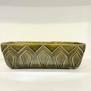 May include: A rectangular, olive green ceramic planter with a geometric diamond pattern. The planter has a glossy finish and is elevated slightly off the surface.