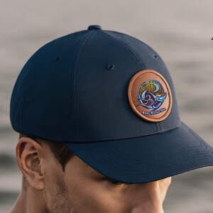 May include: A navy blue baseball cap with a brown leather patch featuring a colorful wave design and the text "Keep Round Sol".