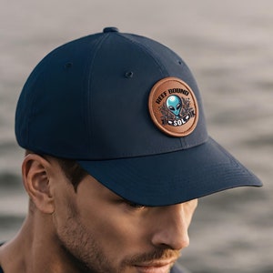 Op de afbeelding: Een marineblauwe baseballpet met een bruin leren patch met een alien-afbeelding en de tekst "Reef Bound Sol".