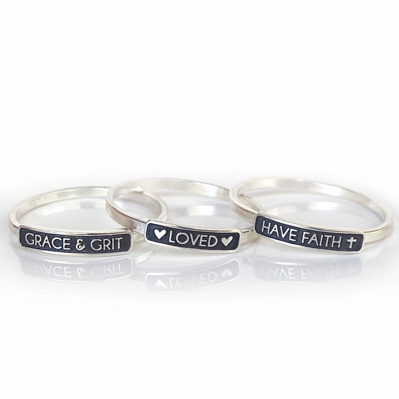 Against the white backdrop we have to the left the GRACE & GRIT plate ring, in the middle we have the silver heart LOVED silver heart plate ring and lastly the HAVE FAITH silver cross plate ring.
