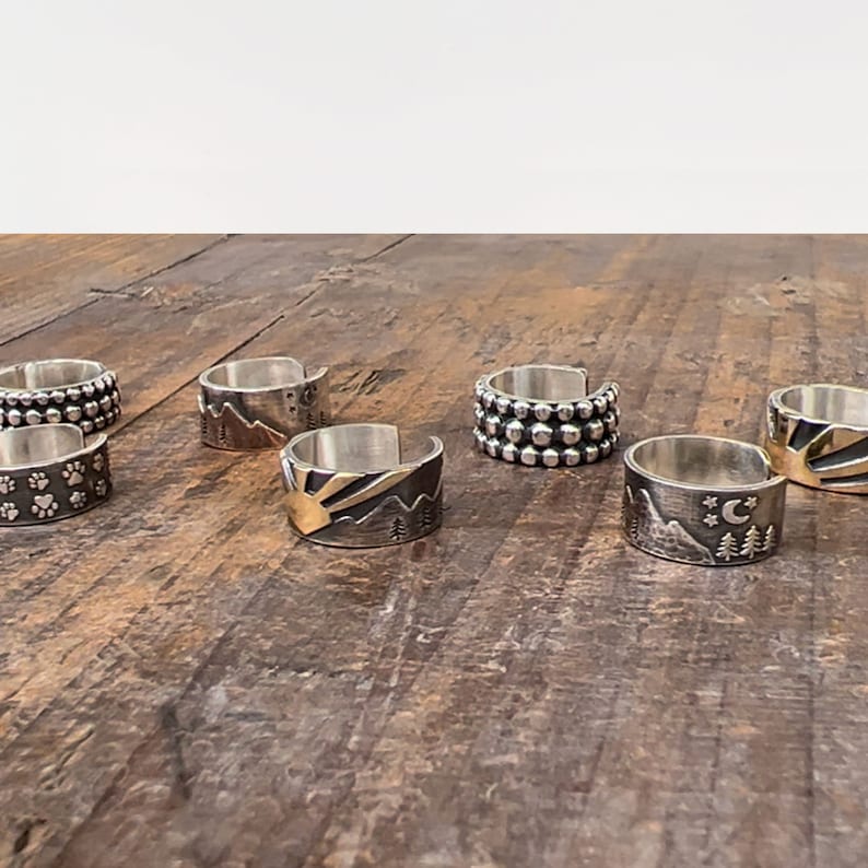 Puede incluir: Un conjunto de seis anillos de plata con diferentes dise&ntilde;os. Los anillos presentan una variedad de patrones, incluyendo huellas de patas, monta&ntilde;as y un sol y una luna. Los anillos son todos de plata y tienen un acabado pulido.