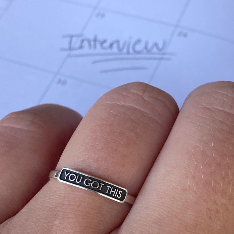 May include: A silver ring with a black band that says "YOU GOT THIS" on it. The ring is worn on a person's finger.