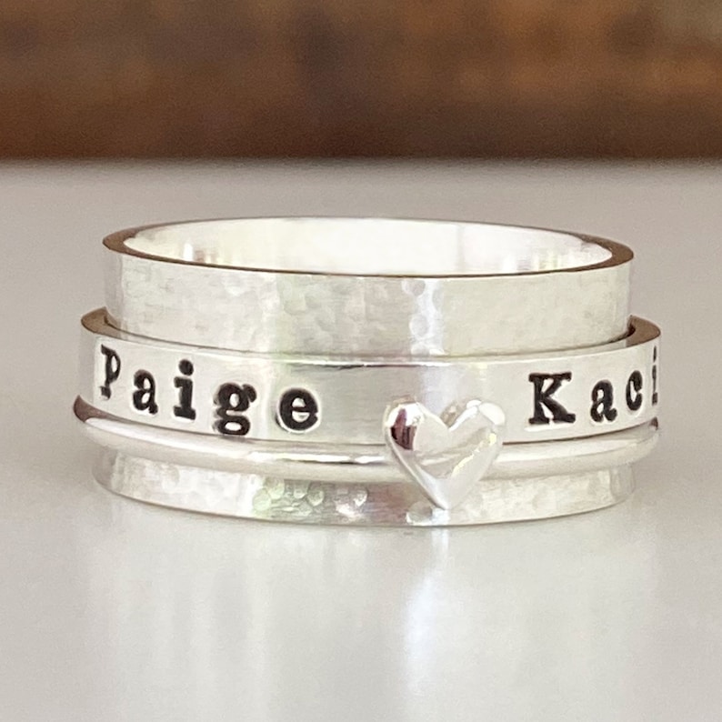 May include: Silver spinning ring engraved with the names "Paige" and "Kaci", separated by a heart charm.