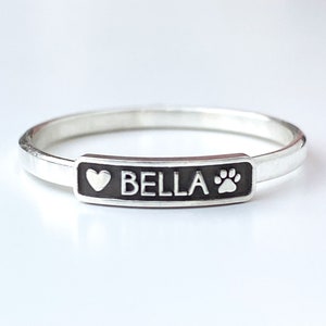 Puede incluir: Anillo de plata con una etiqueta rectangular negra que dice "BELLA" con un corazón y una huella de pata a cada lado.