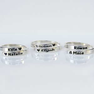 May include: Three silver rings with engraved names and hearts. The rings are stacked on top of each other. The names on the rings are Kyle & Natalie, Vivian & Elijah, and Rowan & Mace.