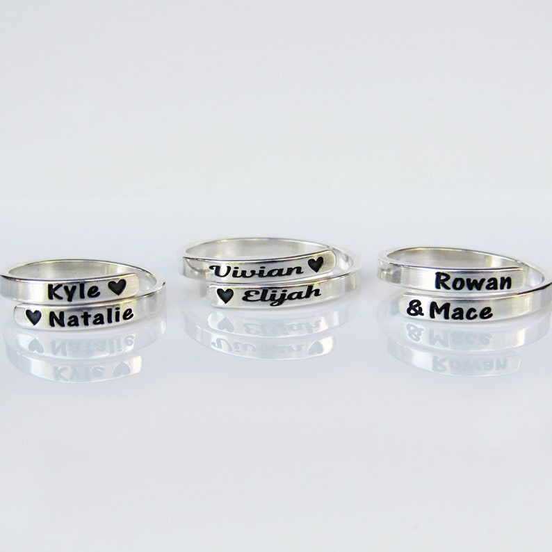 May include: Three silver rings with engraved names and hearts. The rings are stacked on top of each other. The names on the rings are Kyle & Natalie, Vivian & Elijah, and Rowan & Mace.