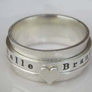 May include: A silver spinning ring with the words "Belle" and "Brand" engraved on the band. A heart-shaped charm is centered on the ring.