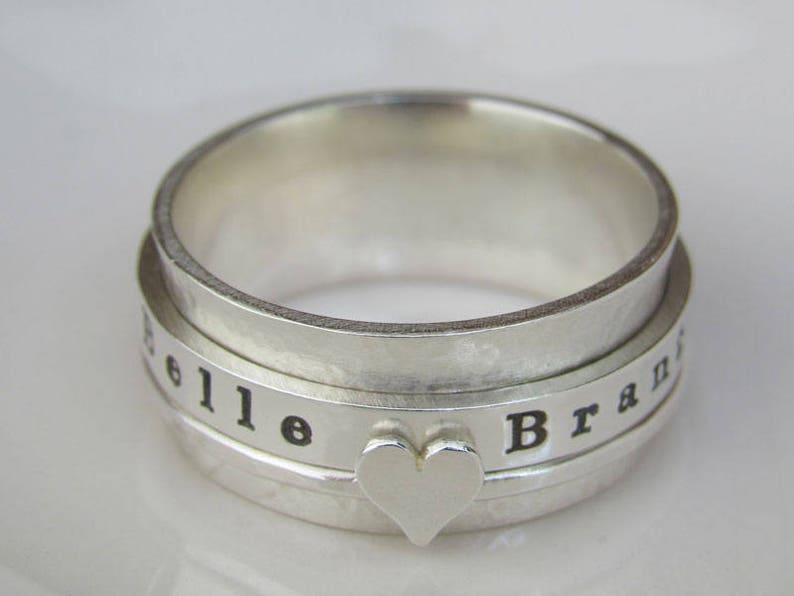 May include: A silver spinning ring with the words "Belle" and "Brand" engraved on the band. A heart-shaped charm is centered on the ring.