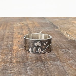 Puede incluir: Un anillo de plata con una escena de monta&ntilde;a grabada. La escena incluye un sol, &aacute;rboles y un p&aacute;jaro volando sobre las monta&ntilde;as.