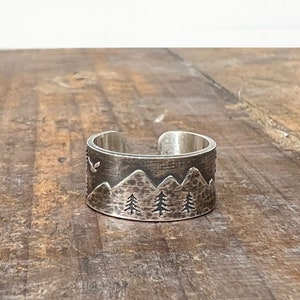 Puede incluir: Un anillo de plata con textura martillada presenta una cordillera y tres pinos. Un p&aacute;jaro vuela sobre las monta&ntilde;as.