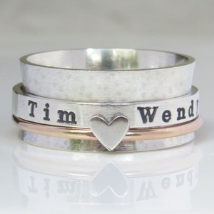 Puede incluir: Un anillo giratorio de plata con los nombres "Tim" y "Wendy" grabados. El anillo tiene un pequeño dije en forma de corazón en el centro.