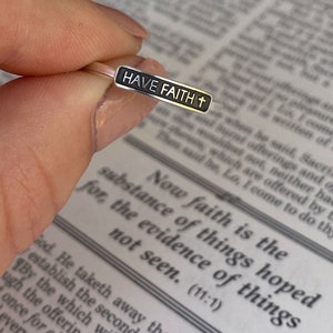 May include: A silver ring with a black rectangular band that says "HAVE FAITH" with a small cross symbol. The ring is on a finger over a page of a book.