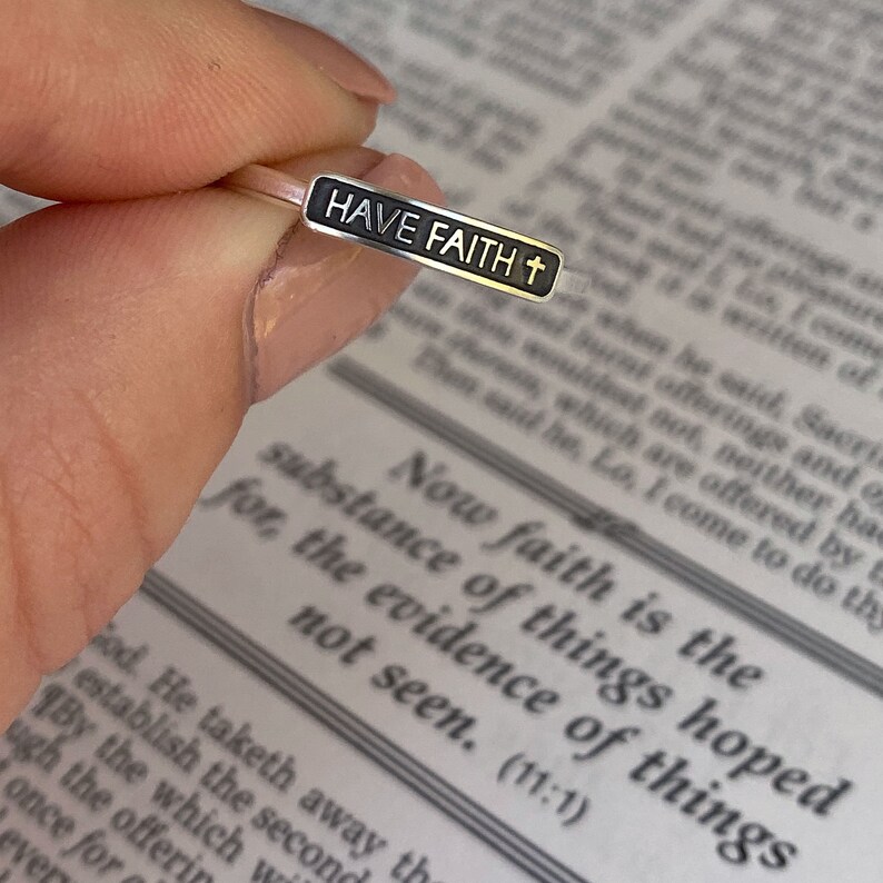 May include: A silver ring with a black rectangular band that says "HAVE FAITH" with a small cross symbol. The ring is on a finger over a page of a book.