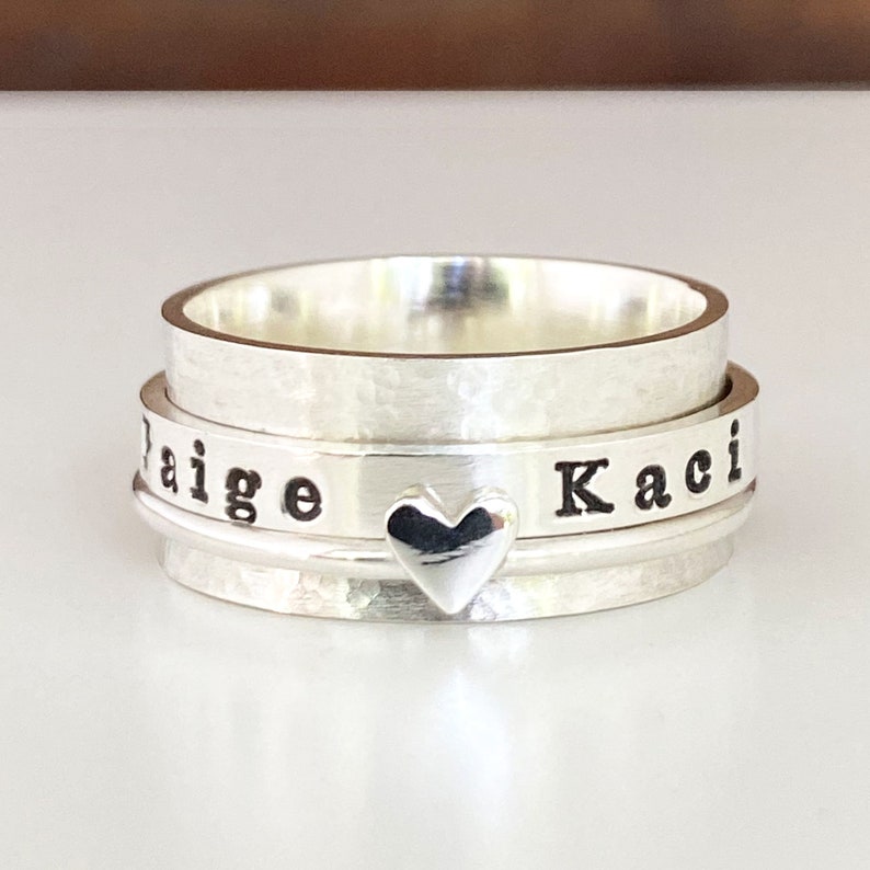 May include: Silver spinning ring engraved with the names "Kaci" and "Paige", separated by a small heart charm.