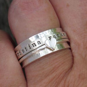 May include: A silver ring with three bands, each engraved with a name. The top band reads "rachel", the middle band reads "christina", and the bottom band has a small heart charm.