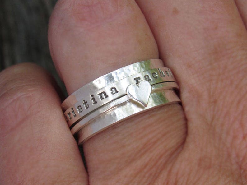 May include: A silver ring with three bands, each engraved with a name. The top band reads "rachel", the middle band reads "christina", and the bottom band has a small heart charm.