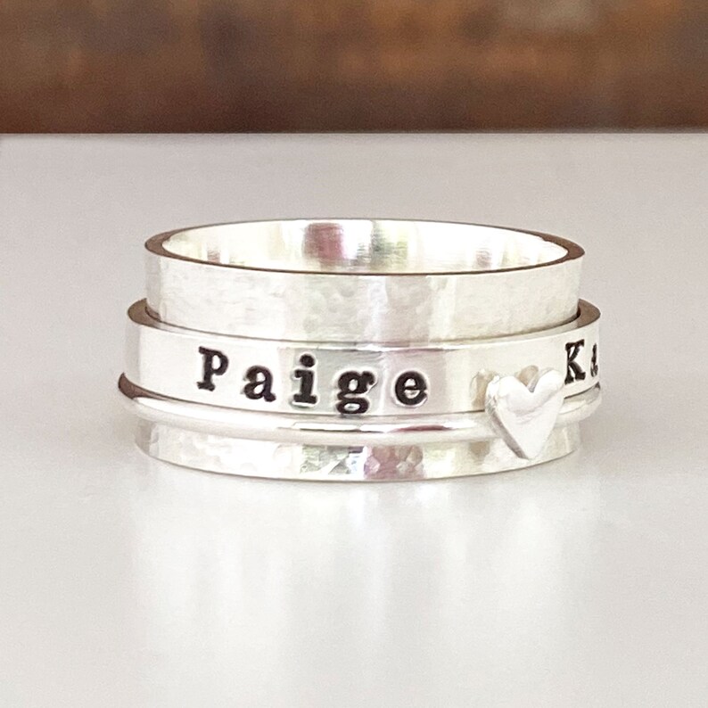 May include: A silver spinning ring with the name "Paige" engraved on one band and a small heart charm on another band.
