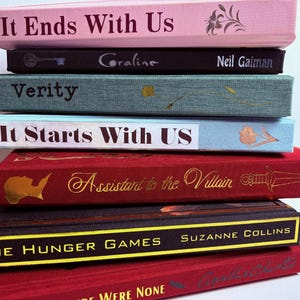 Pode incluir: Uma pilha de livros de capa dura com vários títulos e cores. Os títulos incluem "It Ends With Us", "Coraline", "Verity", "It Starts With Us", "Assistant to the Villain" e "The Hunger Games". Os livros são em tons de rosa, preto, verde-azulado, vermelho e cinza escuro.