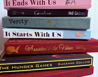 May include: A stack of hardcover books with various titles and colors. The titles include "It Ends With Us," "Coraline," "Verity," "It Starts With Us," "Assistant to the Villain," and "The Hunger Games." The books are in shades of pink, black, teal, red, and dark grey.