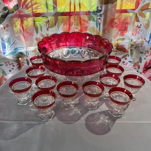 May include: A large glass punch bowl with a scalloped rim and a deep red band, accompanied by twelve matching glass punch cups. Each piece features a clear glass base with a repeating circular pattern and a vibrant red rim around the top edge.
