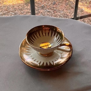 May include: A vintage teacup and saucer set. The cup and saucer are white with gold trim and a repeating geometric pattern. The interior of the cup is gold. The handle is white with gold accents. A gold coin sits inside the cup.