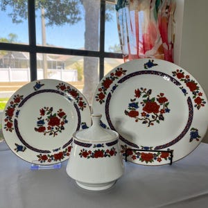 May include: A white porcelain dish set featuring red and blue floral designs. The set includes two plates and a lidded sugar bowl. The plates have a gold trim and a dark blue border with a repeating pattern. The sugar bowl has a matching floral design.