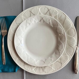 May include: A white ceramic dinner plate and a smaller salad plate, both featuring a raised shell pattern along the rim. The set includes two silver forks resting on a teal napkin, plus a silver dinner knife and a silver spoon placed to the right.