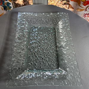 May include: A rectangular clear glass tray featuring a textured, pebbled surface pattern. The tray has a smooth, flat central area surrounded by a raised, wider border with a distinct, uneven, and bumpy finish that catches the light.