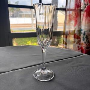 May include: A clear, elegant champagne flute with a long stem and a wide, round base. The glass features vertical ribbed detailing on the upper portion and a diamond-patterned design near the base of the bowl. The glass is set on a gray surface.
