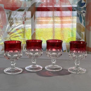 May include: A set of four stemmed glasses featuring a deep red upper bowl and a clear, textured glass base with a repeating circular pattern. Each glass has a short, sturdy stem and a wide, circular foot, showcasing a clear contrast between the colored rim and the patterned lower section of the vessel.