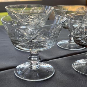 May include: A set of clear crystal stemmed dessert bowls. Each bowl features intricate starburst and geometric patterns. The bowls have a rounded base and a decorative stem. The bowls are arranged on a dark gray surface.