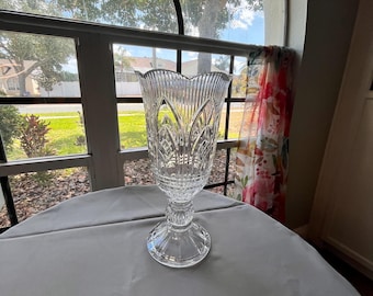 May include: A clear, ornate crystal vase with a wide, scalloped rim and a pedestal base. The vase features vertical and patterned cuts, reflecting light. It sits on a gray tablecloth near a window with a view of a residential area.