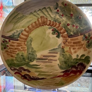 May include: A round, hand-painted ceramic plate featuring a garden scene. The design includes a brick archway, steps, and lush greenery with red berries. The color palette is soft, with earth tones and pastel shades.