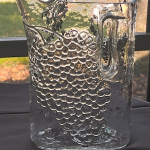 May include: A clear glass pitcher with a textured grape design. The pitcher has a handle and a wide mouth.
