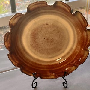 May include: A round ceramic pie dish with a ruffled, scalloped edge. The dish features a gradient glaze that transitions from a light, sandy beige center to a deep, glossy brown rim. The surface has a smooth, reflective finish with subtle texture.