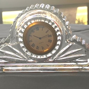 May include: A clear, crystal quartz clock with a gold-colored clock face and Roman numerals. The clock has a decorative, geometric design and is set on a gray surface. The clock is a decorative item.