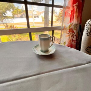 May include: A vintage teacup and saucer set. The cup is white with a light blue rim and handle, and the saucer has a scalloped edge with a matching blue trim. The set is placed on a gray tablecloth near a window.
