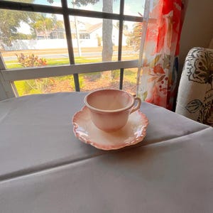 May include: A ceramic teacup and saucer set featuring a soft pink and white gradient glaze. The teacup has a rounded body with a small handle, while the saucer displays a scalloped, wavy edge with a matching pink and white color pattern.