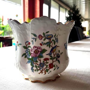 May include: White ceramic planter with a scalloped rim and gold trim. The planter is decorated with colorful floral and bird motifs. The flowers are pink, blue, and red, with green leaves. A small bird perches on a branch.