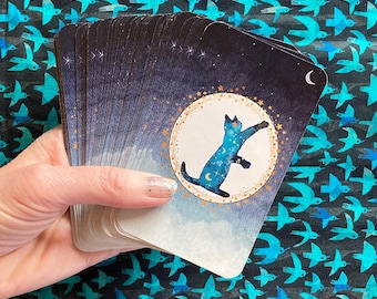 May include: A deck of cards with a blue cat jumping in a circle of stars and a crescent moon on the top card. The cards have a dark blue background with gold accents.