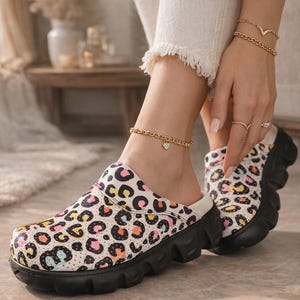 May include: Clogs with a colorful leopard print pattern in pink, orange, yellow, and teal on a white background. The clogs have a black sole and a white interior. A gold anklet with a heart charm is visible.