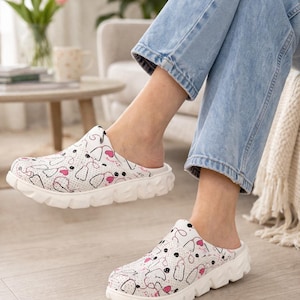 Puede incluir: Zuecos blancos sin cordones con un estampado lúdico de puntos negros, corazones rosas y dibujos lineales. Los zuecos tienen una suela blanca gruesa y texturizada. La imagen muestra los zuecos con vaqueros azules.