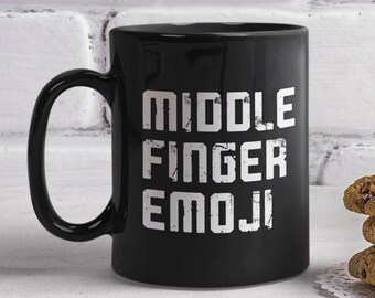 May include: Black coffee mug with white text that reads "MIDDLE FINGER EMOJI". The mug is sitting on a white surface with a stack of chocolate chip cookies and a white vase with a yellow orchid.