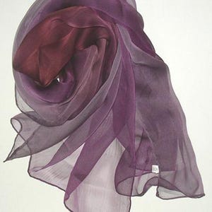 May include: A sheer purple and burgundy ombre scarf made of silk. The scarf is draped and folded, showing the gradient of colour.
