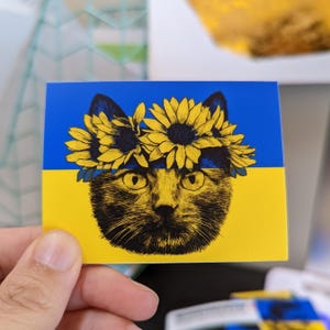 May include: A black cat with a sunflower crown on a blue and yellow background. The colors of the flag of Ukraine.