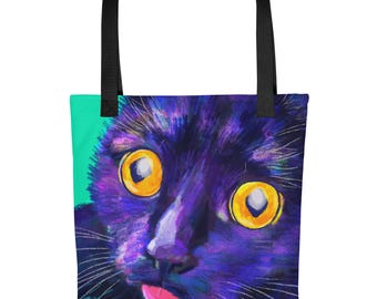 May include: A tote bag with black handles featuring a vibrant, painterly illustration of a black cat with large, bright yellow eyes and a small pink tongue sticking out, set against a deep purple and blue background with a teal side panel.