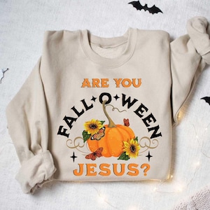 May include: A beige sweatshirt with the text "Are you Fall-o-ween Jesus?" printed on it. The text is surrounded by a pumpkin, sunflowers, and butterflies.