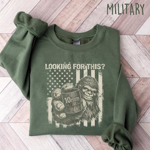May include: Military green sweatshirt with the text "LOOKING FOR THIS?" above a graphic of the American flag and a Bigfoot holding a 10mm cartridge. The sweatshirt is folded on a light wood-grain surface.