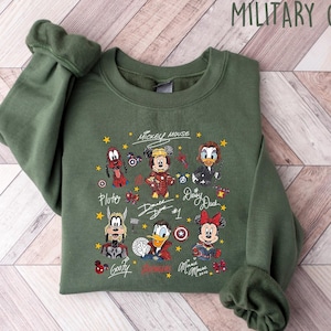 May include: Military green sweatshirt featuring a colorful graphic of Mickey Mouse and friends, including Pluto, Donald Duck, and Minnie Mouse. The design includes character signatures and the text "Mickey Mouse". The sweatshirt is made of a soft material.